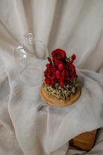 Load image into Gallery viewer, Decorative arrangement of red roses and greenery on a wooden base with a glass cover, placed on a white fabric background.
