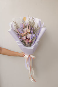 Bouquet of flowers wrapped in purple paper with a ribbon, held by a person against a plain background