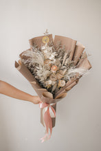 Load image into Gallery viewer, Bouquet of dried flowers wrapped in brown paper with a pink ribbon, held by a hand against a plain background.
