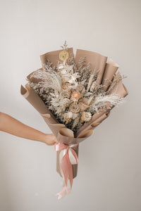 Bouquet of dried flowers wrapped in brown paper with a pink ribbon, held by a hand against a plain background.