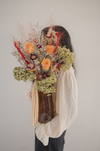 Person holding a large, ornate vase of dried and preserved flowers against a plain background.