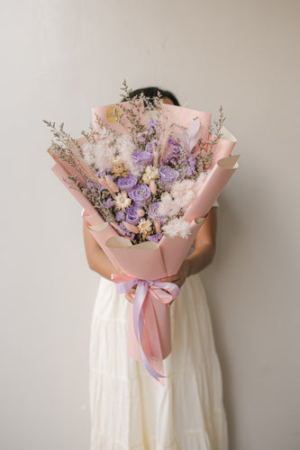 Person holding a bouquet of flowers wrapped in pink paper against a plain background