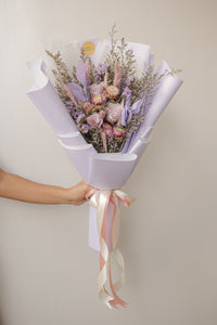 Bouquet of flowers wrapped in purple paper with a ribbon, held by a person against a plain background.