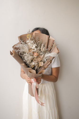 Person holding a bouquet of dried flowers wrapped in brown paper against a plain background