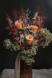 TS12 inspired dried flowers with orange roses and greenery in a clear vase against a dark background.
