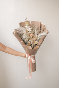 Bouquet of dried flowers wrapped in brown paper with a pink ribbon, held against a plain background.